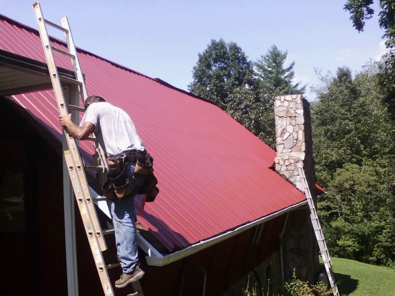 Roofer on a ladder beside a red metal roof and stone chimney — Soffit & Fascia Repair by Roofing Brothers in Dazey
